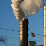 Industrial Smokestacks Releasing Pollution – Asbestos Exposure Context - Ferraro Law Firm Factory smokestacks emitting thick white smoke with an American flag visible below, symbolizing industrial asbestos exposure and related lawsuits.