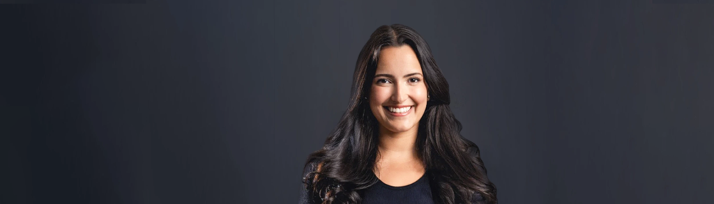 Smiling woman with long dark hair, wearing a dark top, head-and-shoulders portrait against a dark gray gradient background.