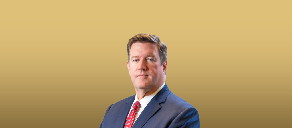 Portrait of a man in a navy suit and red tie against a gold gradient background, looking confident.