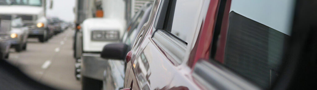 Close-up of a red car in heavy traffic, with trucks and other vehicles lined up in the distance.