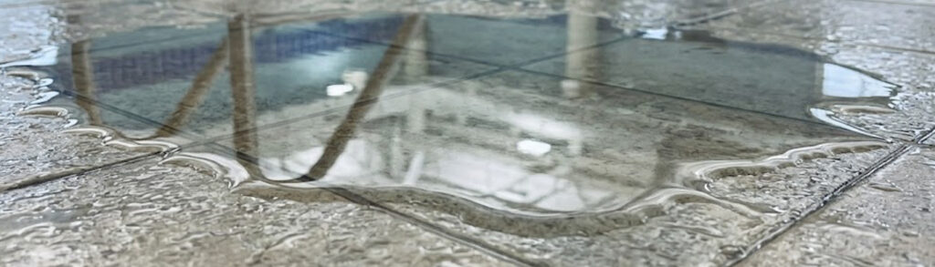 Puddle on wet stone pavement reflecting nearby buildings and sky beyond the edge of the crack.