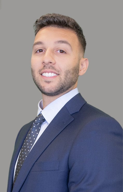 Professional headshot of a smiling man wearing a navy suit, white shirt, and dotted tie against a gray backdrop