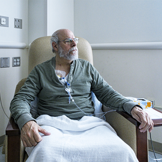 Older man with gray beard and glasses sits in a hospital chair, connected to tubing and covered with a white blanket.