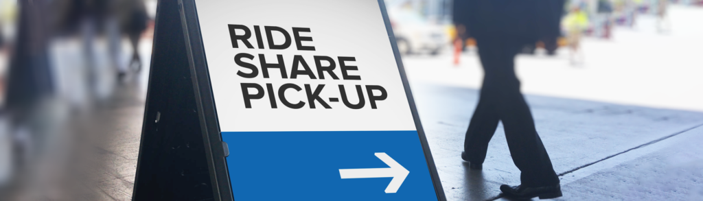 Sign reading 'RIDE SHARE PICK-UP' with a blue bottom and a white right-facing arrow on a sidewalk.