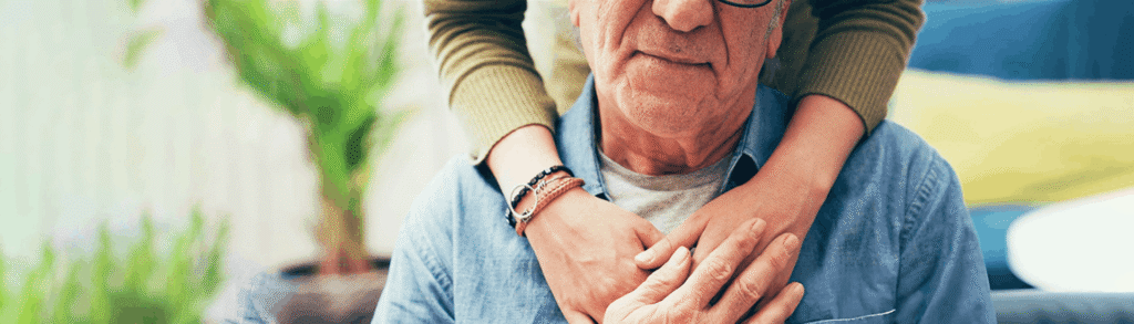 Older man in a denim shirt being hugged from behind by a younger person.