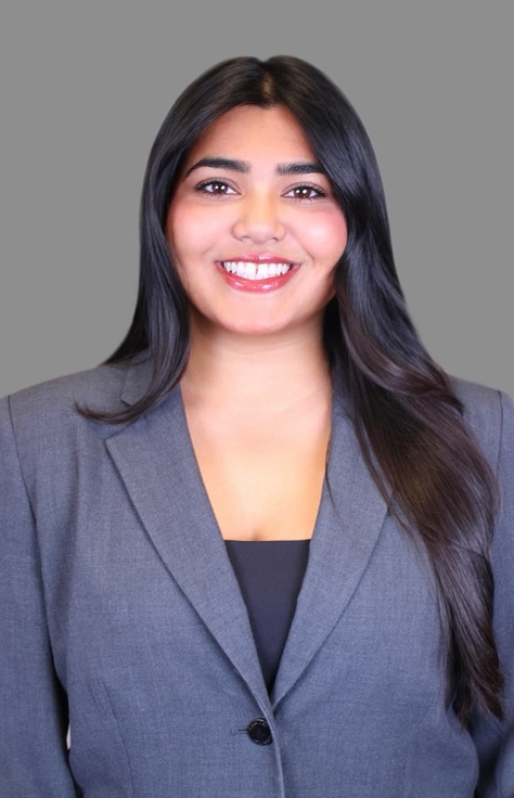 Professional headshot of Brhea D’Mello, a smiling woman with long dark hair wearing a gray blazer against a gray backdrop